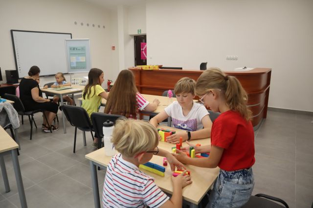 dzieci siedzą przy brązowych stołach i graja w gry planszowe, za nimi plakat z informacjami na temat projektu BLISKO