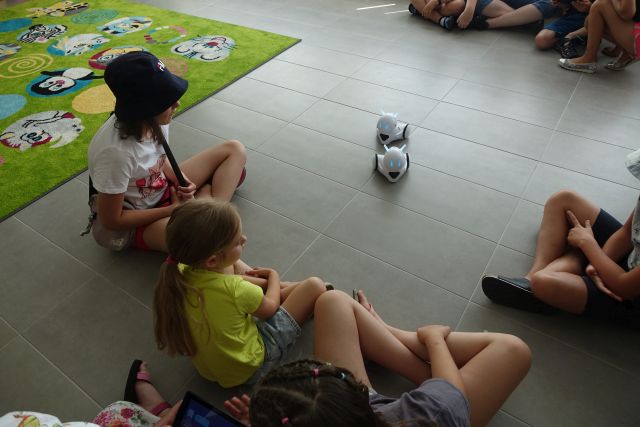 na siwej podłodze siedzą dzieci, przed nimi dwa białe małe roboty Photon