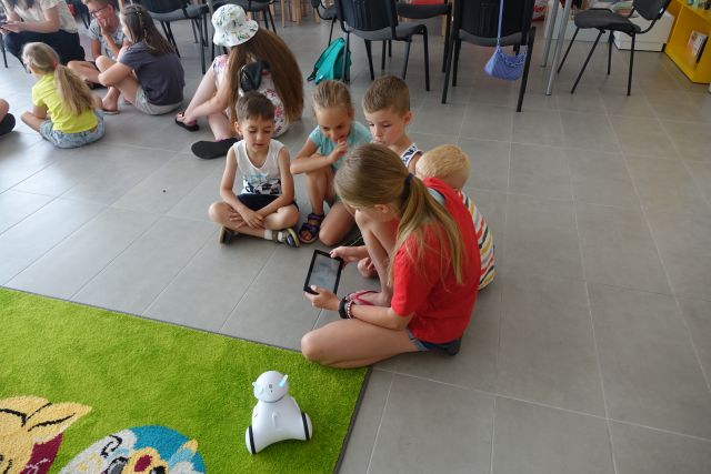 dzieci siedzą na siwej podłodze, dziewczynka trzyma w dłoniach tablet, na zielonym dywanie stoi mały robot biały Photon