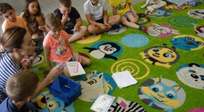 na zielonym dywanie siedzą dzieci i kobieta, przed nią niebieski karton i kartki z liczbami