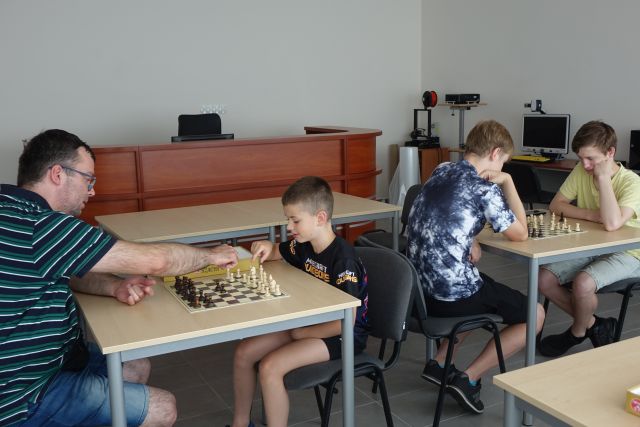 dzieci i młodzież oraz trener siedzą przy brązowych stołach i grają w szachy, za nimi brązowe biurko