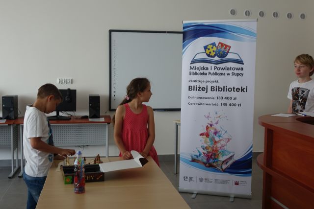 chłopiec siedzi przy brązowym stole i rozkłada planszę z szachami, dziewczynka stoi, w drzwiach stoi chłopiec, za nimi duży plakat z nazwą zadania i logo biblioteki