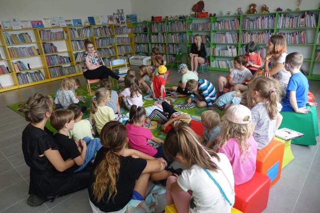 na zielonym dywanie siedzą dzieci, na czerwonych pufach siedzą kobiety, za nimi zielone i żółte regały z książkami
