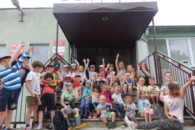 liczna grupa dzieci i opiekunów stoi na schodach biblioteki, pod zadaszeniem, ręce trzymają uniesione do góry