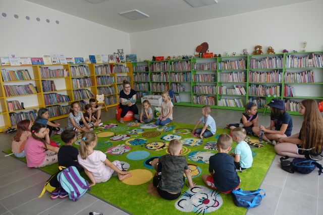 dzieci i kobieta czytająca książkę siedzą na zielonym dywanie, za nimi żółte i zielone regały z książkami