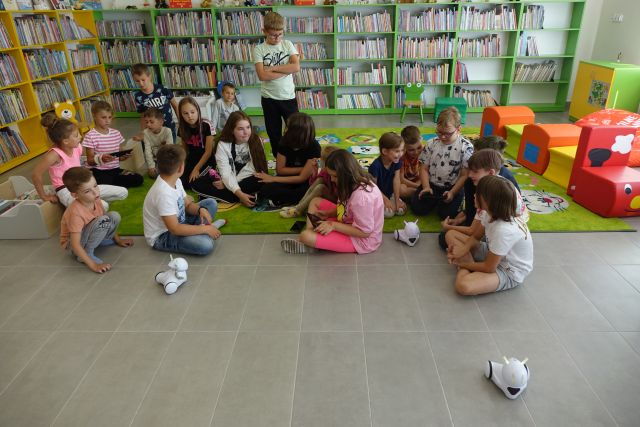 dzieci siedzą na zielonym dywanie i na siwej podłodze, przed nimi trzy małe białe roboty, za ni mi żółte i zielone regały z książkami