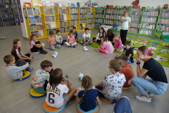dzieci siedzą w kole na siwej podłodze, przed nimi białe małe roboty, za nimi zielone i żółte regały z książkami