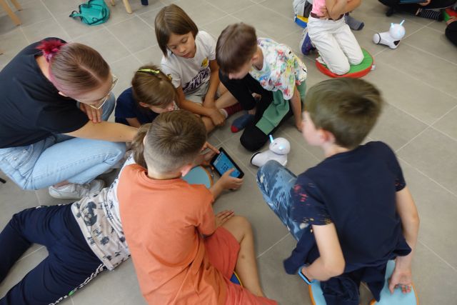 na siwej podłodze siedzą dzieci, chłopiec w dłoniach trzyma tablet, w środku na podłodze biały mały robot