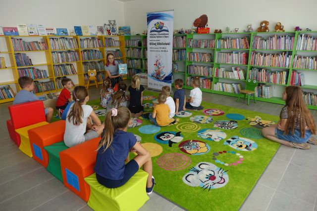na zielonym dywanie siedzą dzieci, przed nimi kobieta siedzi na krześle, w dłoniach ma książkę, za nią plakat z logo biblioteki na rozłożonej książce herb Słupcy na niebieskim tle żółty krzyż, herb powiatu słupeckiego na czerwono niebieskim tle biały orzeł, nazwa zadania, w tle zielone i żółte regały z książkami