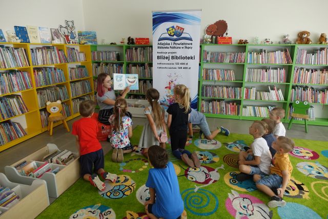 na zielonym dywanie siedzą dzieci, przed nimi kobieta siedzi na krześle, w dłoniach ma książkę, za nią plakat z logo biblioteki na rozłożonej książce herb Słupcy na niebieskim tle żółty krzyż, herb powiatu słupeckiego na czerwono niebieskim tle biały orzeł, nazwa zadania, w tle zielone i żółte regały z książkami
