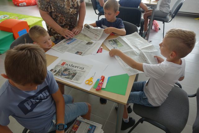 przy brązowym stole siedzą dzieci i wycinają z gazet