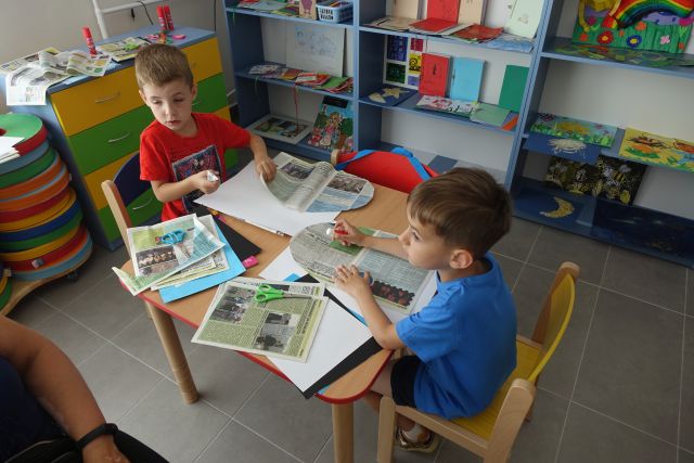 przy brązowym stole siedzą dzieci i wycinają z gazet
