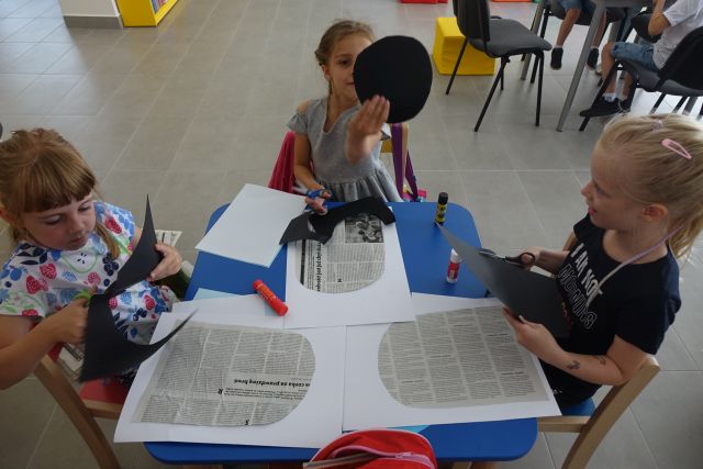 na stole leżą białe i niebieskie kartki oraz wycinki z gazet, dziewczynka trzyma w ręce czarne koło