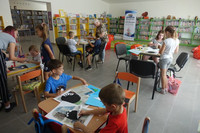 przy stołach siedzą dzieci i naklejają wycięte z kartek zęby rekina, za nimi żółte i zielone regały z książkami