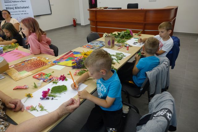 Przy brązowych stołach siedzą dzieci i wyklejają marzenia z kwiatów i traw