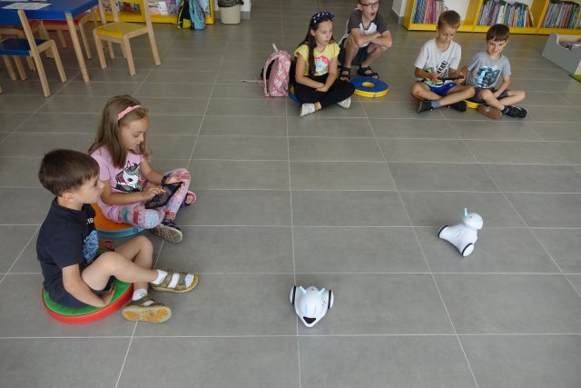 na siwej podłodze siedzą dzieci i jeżdżą małe białe roboty