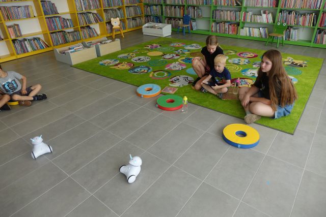 na zielonym dywanie siedzą dzieci, po podłodze jeżdżą małe białe roboty