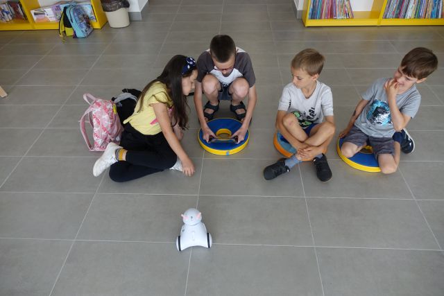 na podłodze siedzą dzieci, chłopiec w dłoniach trzyma tablet, przed nimi jeździ biały mały robot