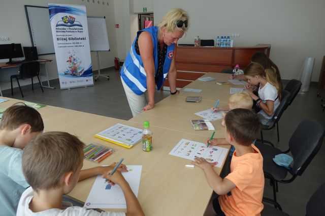 przy brązowych stołach siedzą dzieci, wypełniają krzyżówki, przed nimi stoi oparta o stół kobieta