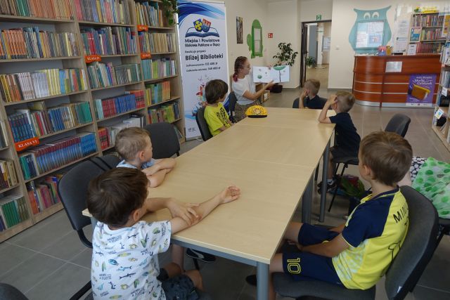 przy brązowych stołach siedzą dzieci , na końcu stołu siedzi kobieta i trzyma książkę, z jej lewej strony stoi duży plakat z logo biblioteki na rozłożonej książce herb Słupcy - na niebieskim tle żółty krzyż oraz herb powiatu słupeckiego - czerwono-niebieskie tło, na czerwonym tle biały orzeł, na niebieskim tle żółte krzyże, z lewe strony brązowe regały z książkami