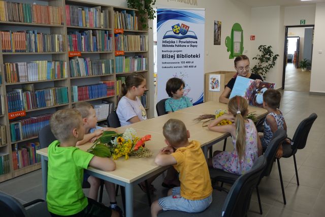 dzieci siedzą przy brązowych stołach, kobieta siedzi na końcu stołu itrzyma w rękach książkę, z lewej strony regały z książkami, z lewej strony kobiety plakat z informacją o projekcie i logo biblioteki - na rozłożonej książce herb miasta na niebieskim tle żółty krzyż, oraz herb powiatu słupeckiego, od góry na czerwonym tle biały orzeł do połowy, od dołu na niebieskim tle wokoło małe żółte krzyże