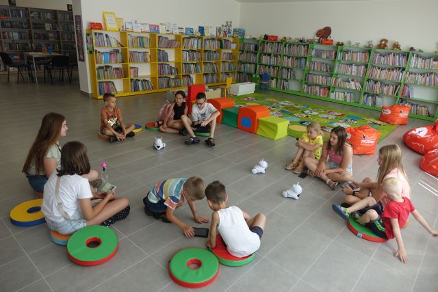 na podłodze z siwych kafli siedzą dzieci na kolorowych pufach, przed nimi małe białe roboty, za nimi zielony dywan i zielone i żółte regały z książkami