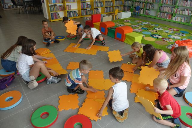 dzieci na siwej podłodze układają duże pomarańczowe puzle, za nimi zielone, czerwone i żółte pufy tworzące pociąg oraz zielony dywan i zielone i żółte regały z książkami