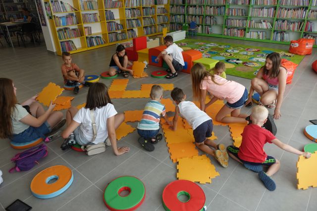 dzieci na siwej podłodze układają duże pomarańczowe puzle, za nimi zielone, czerwone i żółte pufy tworzące pociąg oraz zielony dywan i zielone i żółte regały z książkami