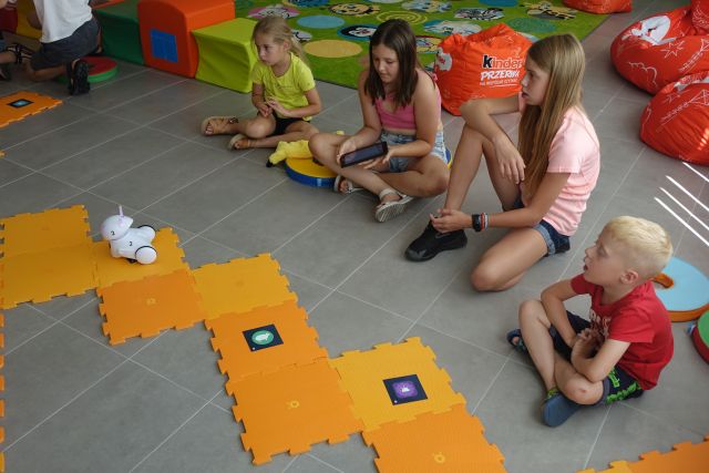 dzieci siedzą na kolorowych pufkach na podłodze przed nimi pomarańczowe duże puzle, a po nich jeżdżą roboty