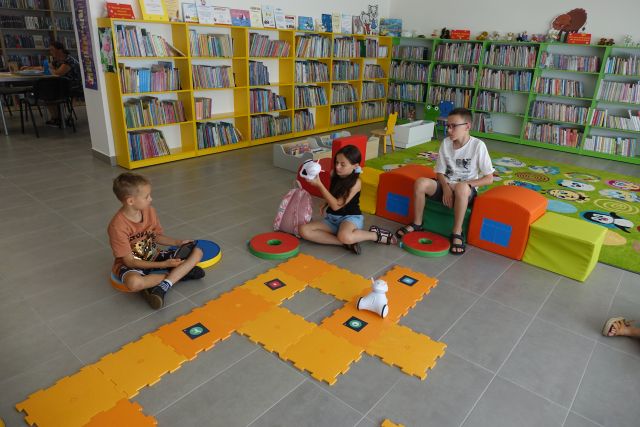 dzieci siedzą na kolorowych pufkach na podłodze przed nimi pomarańczowe duże puzle, a po nich jeżdżą roboty