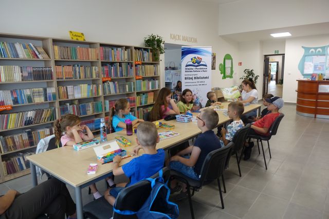 przy brązowych stołach siedzą dzieci, na końcu siedzi kobieta i czyta książkę, za dziećmi brązowe regały z książkami, za kobietą duży plakat z informacja o dofinansowaniu i logo biblioteki - na rozłożonej książce herb Słupcy na niebieskim tle żółty krzyż oraz herb powiatu słupeckiego oraz herb powiatu słupeckiego, od góry na czerwonym tle biały orzeł do połowy, od dołu na niebieskim tle wokoło małe żółte krzyże