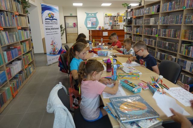 dzieci siedzą przy brązowych stołach i wypełniają krzyżówki oraz kolorują kolorowanki, przed nimi plakat z informacja o dofinansowaniu z logo biblioteki na rozłożonej książce herb Słupcy na niebieskim tle żółty krzyż oraz herb powiatu słupeckiego, od góry na czerwonym tle biały orzeł do połowy, od dołu na niebieskim tle wokoło małe żółte krzyże