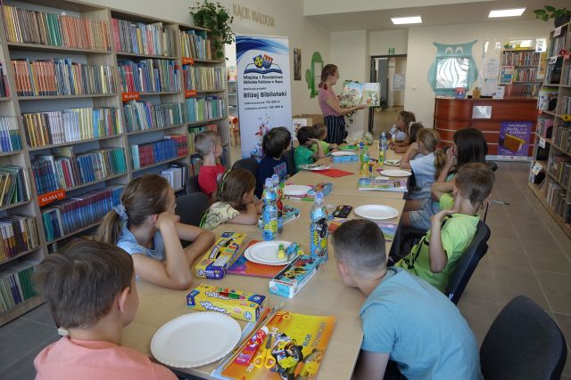 przy brązowych stołach siedzą dzieci, na stołach paierowe talerzyki, farbki, woda w butelkach, przed dziećmi stoi kobieta i pokazuje książkę, za dziećmi brązowe regały z książkami, za kobietą duży plakat z informacja o dofinansowaniu i logo biblioteki - na rozłożonej książce herb Słupcy na niebieskim tle żółty krzyż oraz herb powiatu słupeckiego oraz herb powiatu słupeckiego, od góry na czerwonym tle biały orzeł do połowy, od dołu na niebieskim tle wokoło małe żółte krzyże 