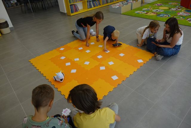 na podłodze rozłożone pomarańczowe  duże puzle, na nich roboty małe białe i dzieci