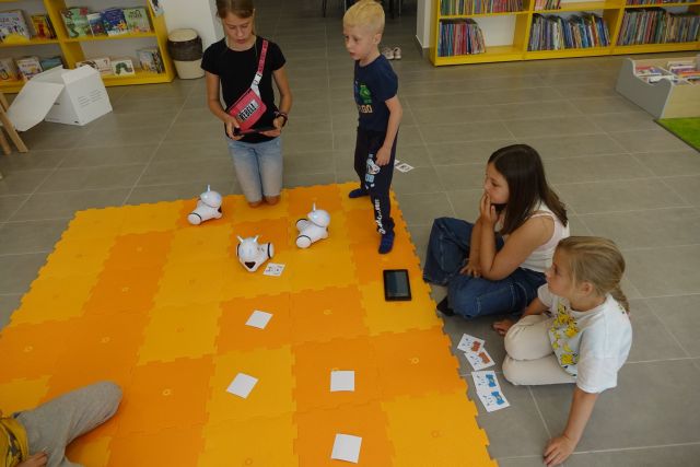 na podłodze rozłożone duże pomaranczowe puzle, na nich małe białe roboty i dzieci