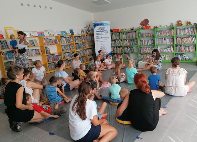 na zielonym dywanie i podłodze siedzą dzieci i osoby dorosłe, przed nimi siedzi kobieta, za nią żółte i zielone regały z książkami, w górnym rogu z lewej strony stoi duży plakat z nazwą projektu i logo biblioteki na rozłożonej książce herb Słupcy na niebieskim tle żółty krzyż oraz herb powiatu słupeckiego oraz herb powiatu słupeckiego, od góry na czerwonym tle biały orzeł do połowy, od dołu na niebieskim tle wokoło małe żółte krzyże