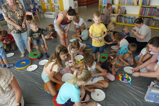 dzieci i dorośli siedzą i stoją na szarej folli, przed sobą mają papierowe talerzyki