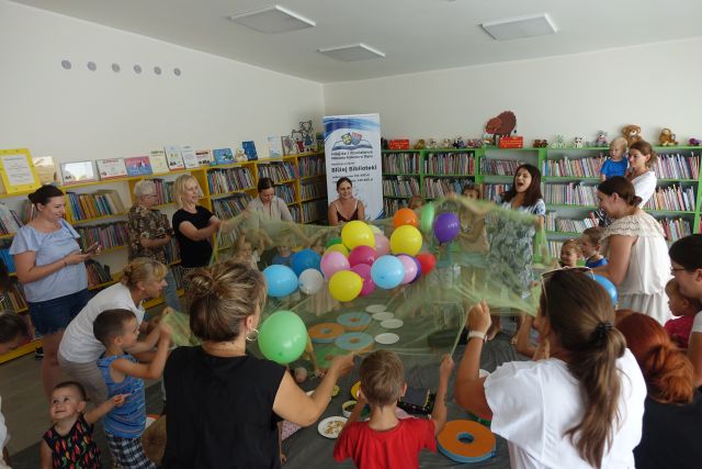 dorośli i dzieci trzymaja materiał i podrzucają na nim balony