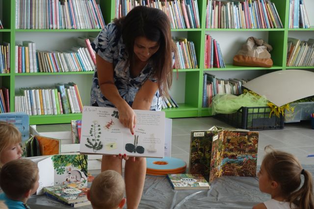 kobie pochylone pokazuje dzieciom siedzącym na zielonym dywanie książkę, za nią zielone regały z książkami