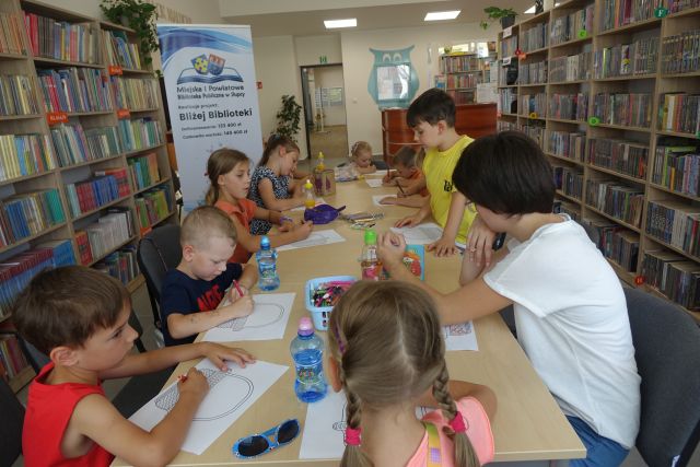 przy brązowym stole siedzą dzieci i malują koszyczki, z lewej strony stoi duży plakat z nazwą projektu i logo biblioteki na rozłożonej książce herb Słupcy na niebieskim tle żółty krzyż oraz herb powiatu słupeckiego oraz herb powiatu słupeckiego, od góry na czerwonym tle biały orzeł do połowy, od dołu na niebieskim tle wokoło małe żółte krzyże