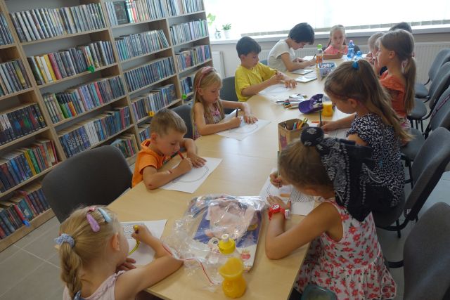 przy brązowych stołach siedzą dzieci i malują koszyczki