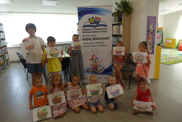 dzieci stoją i prezentują swoje prace : pomalowane koszyczki a w nich owoce i warzywa, za dziećmi stoi duży plakat z nazwą projektu i logo biblioteki na rozłożonej książce herb Słupcy na niebieskim tle żółty krzyż oraz herb powiatu słupeckiego oraz herb powiatu słupeckiego, od góry na czerwonym tle biały orzeł do połowy, od dołu na niebieskim tle wokoło małe żółte krzyże