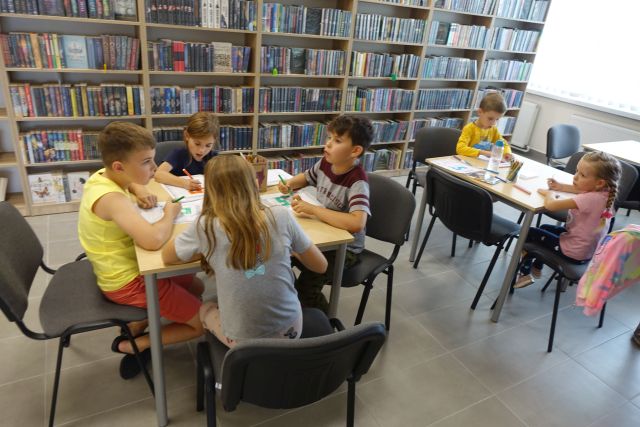 przy brązowych stolikach na siwych krzesłach siedzą dzieci i piszą na kartkach, za nimi brązowe regały z ksiązkami