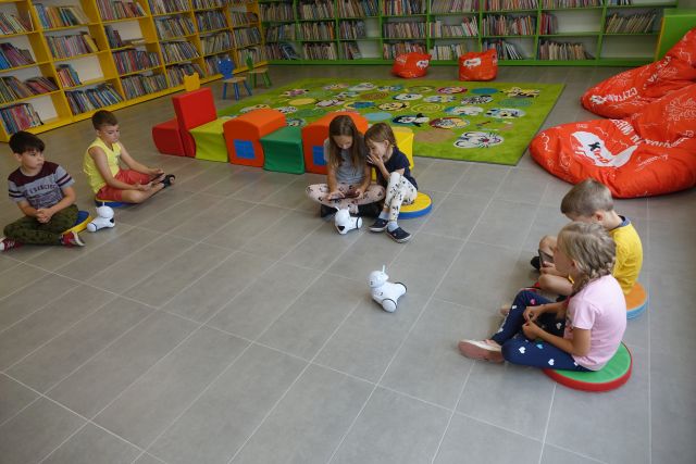 na podłodze siedzą dzieci i sterują małymi białymi robotami, za nimi zielony dywan, kolorowe pufy, za nimi zielone i żółte regały z książkami