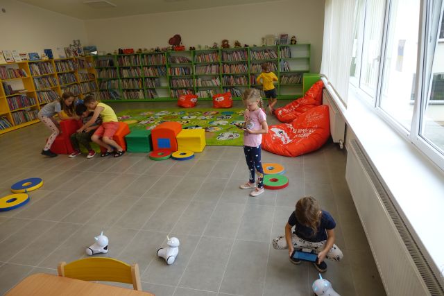 po podłodze jeżdżą małe białe roboty sterowane przez dzieci, z tyłu leży zielony dywan i kolorowe pufy oraz zielone i żółte regały z ksiązkami