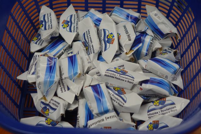 cukierki krówki owinięte w papierki z logo biblioteki na rozłożonej książce herb Słupcy na niebieskim tle żółty krzyż oraz herb powiatu słupeckiego oraz herb powiatu słupeckiego, od góry na czerwonym tle biały orzeł do połowy, od dołu na niebieskim tle wokoło małe żółte krzyże i nazwą projektu