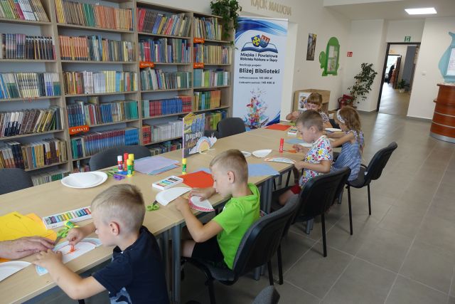 dzieci siedzą przy brązowych stołach i z papierowych talerzyków wykonują dinozaury i je malują, przed nimi stoją regały z książkami, a po prawej strony duży plakat z nazwą projektu i logo biblioteki na rozłożonej książce herb Słupcy na niebieskim tle żółty krzyż oraz herb powiatu słupeckiego oraz herb powiatu słupeckiego, od góry na czerwonym tle biały orzeł do połowy, od dołu na niebieskim tle wokoło małe żółte krzyże