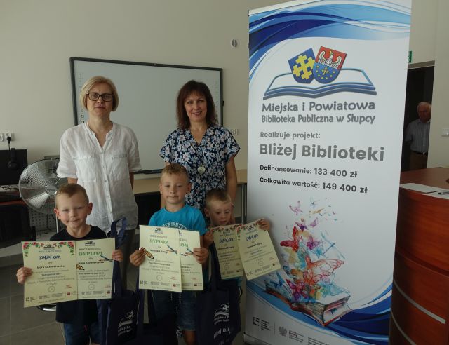 dzieci stoja i trzymaja w rękach dyplomy oraz kolorowe torby, za nimi dwie dorosłe kobiety, z prawej strony stoi duży plakat z nazwą projektu i logo biblioteki na rozłożonej książce herb Słupcy na niebieskim tle żółty krzyż oraz herb powiatu słupeckiego oraz herb powiatu słupeckiego, od góry na czerwonym tle biały orzeł do połowy, od dołu na niebieskim tle wokoło małe żółte krzyże