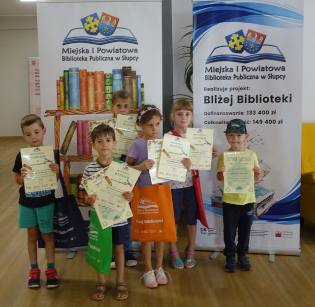 dzieci stoją przed dwoma dużymi plakatami - jeden z narysowanymi książkami i nazwą biblioteki, a drugi z informacja o projekcie i logo biblioteki na rozłożonej książce herb Słupcy na niebieskim tle żółty krzyż oraz herb powiatu słupeckiego oraz herb powiatu słupeckiego, od góry na czerwonym tle biały orzeł do połowy, od dołu na niebieskim tle wokoło małe żółte krzyże. Dzieci trzymają w rękach dyplomy i torby z nazwą biblioteki i projektu