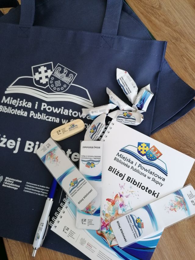 na brązowym stole leżą kolorowe torby z nazwą projektu, zakładki, cukierki, kubek, pendrive